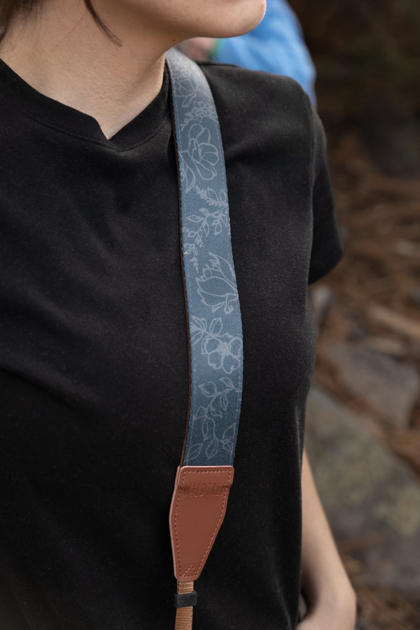 A close-up lifestyle shot of the Shadow Garden camera strap worn over the shoulder. The strap features a moody slate-blue fabric with a delicate light-gray floral pattern, finished with a brown vegan leather end piece embossed with the WILDTREE logo.