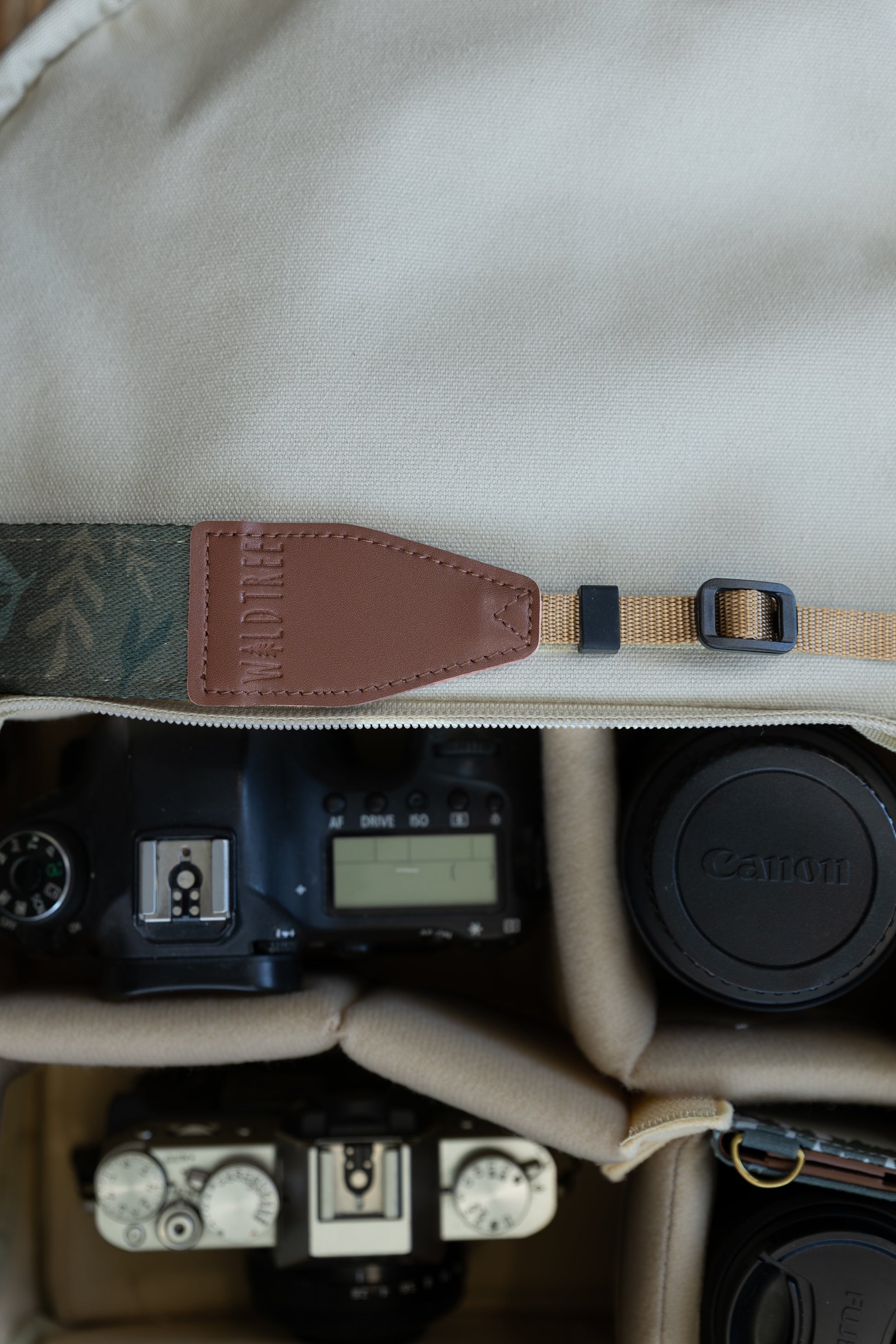 Top-down view of the inside of a camera bag, featuring the WILDTREE "Silent Grove" floral green camera strap resting across the top, above a camera body and lens.