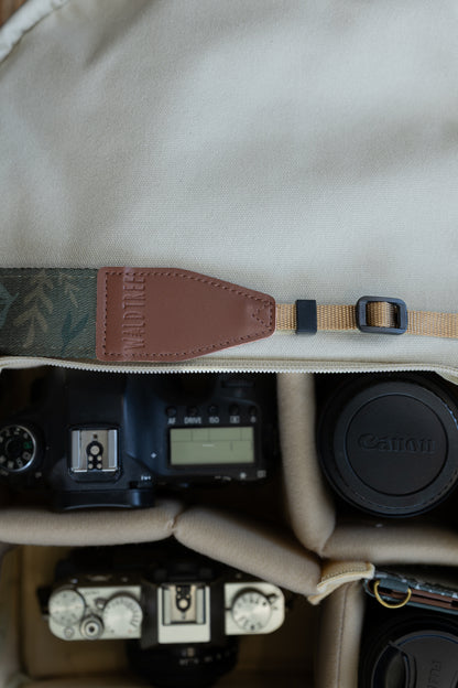 Top-down view of the inside of a camera bag, featuring the WILDTREE "Silent Grove" floral green camera strap resting across the top, above a camera body and lens.