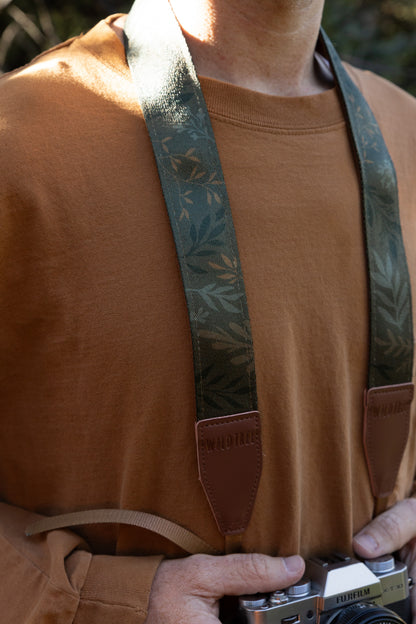 Close-up view of a person wearing the "Silent Grove" green floral camera strap around their neck, with hands holding the Fujifilm camera.
