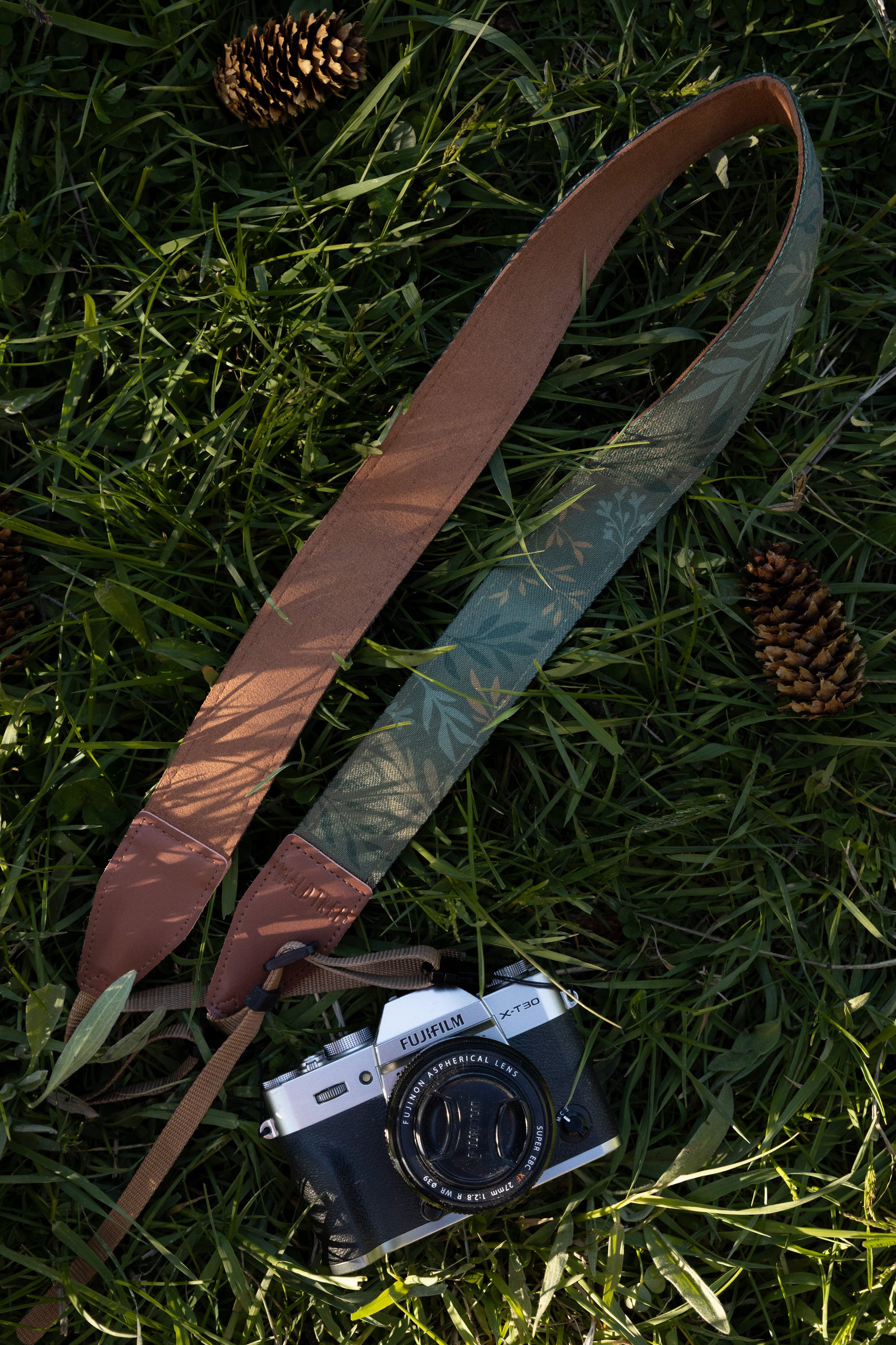 The "Silent Grove" floral camera strap coiled on a grassy outdoor surface next to a Fujifilm camera and pinecone.