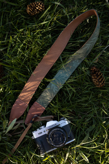 The "Silent Grove" floral camera strap coiled on a grassy outdoor surface next to a Fujifilm camera and pinecone.