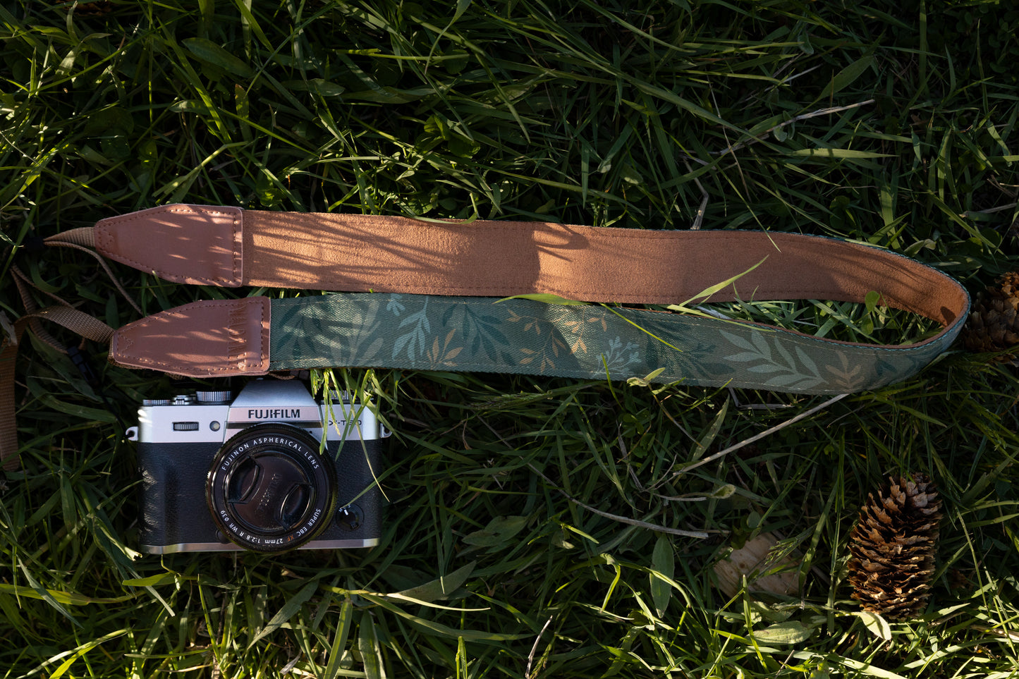 The "Silent Grove" floral camera strap and camera placed on green grass next to a pinecone, showing the strap's length.