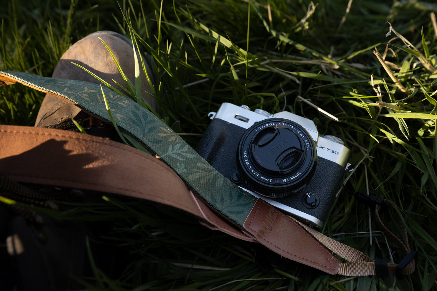 A Fujifilm camera rests in green grass, with the "Silent Grove" green floral strap draped alongside it.