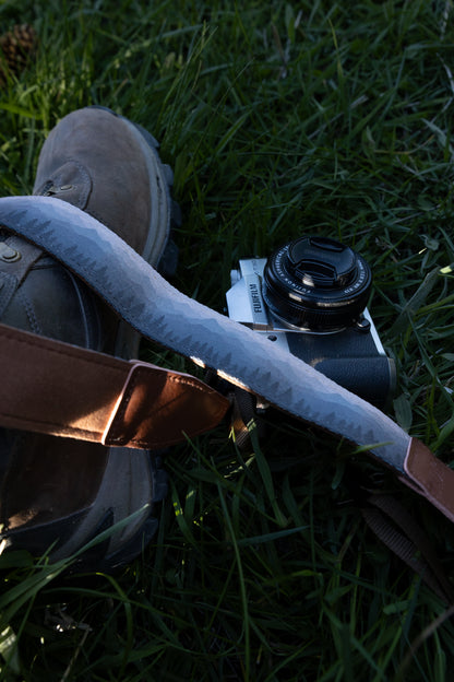 Silent Summit camera strap focusing on the blue-to-gray monochromatic mountain and evergreen forest artwork alongside a hiking boot and vintage-style silver camera on the lawn.