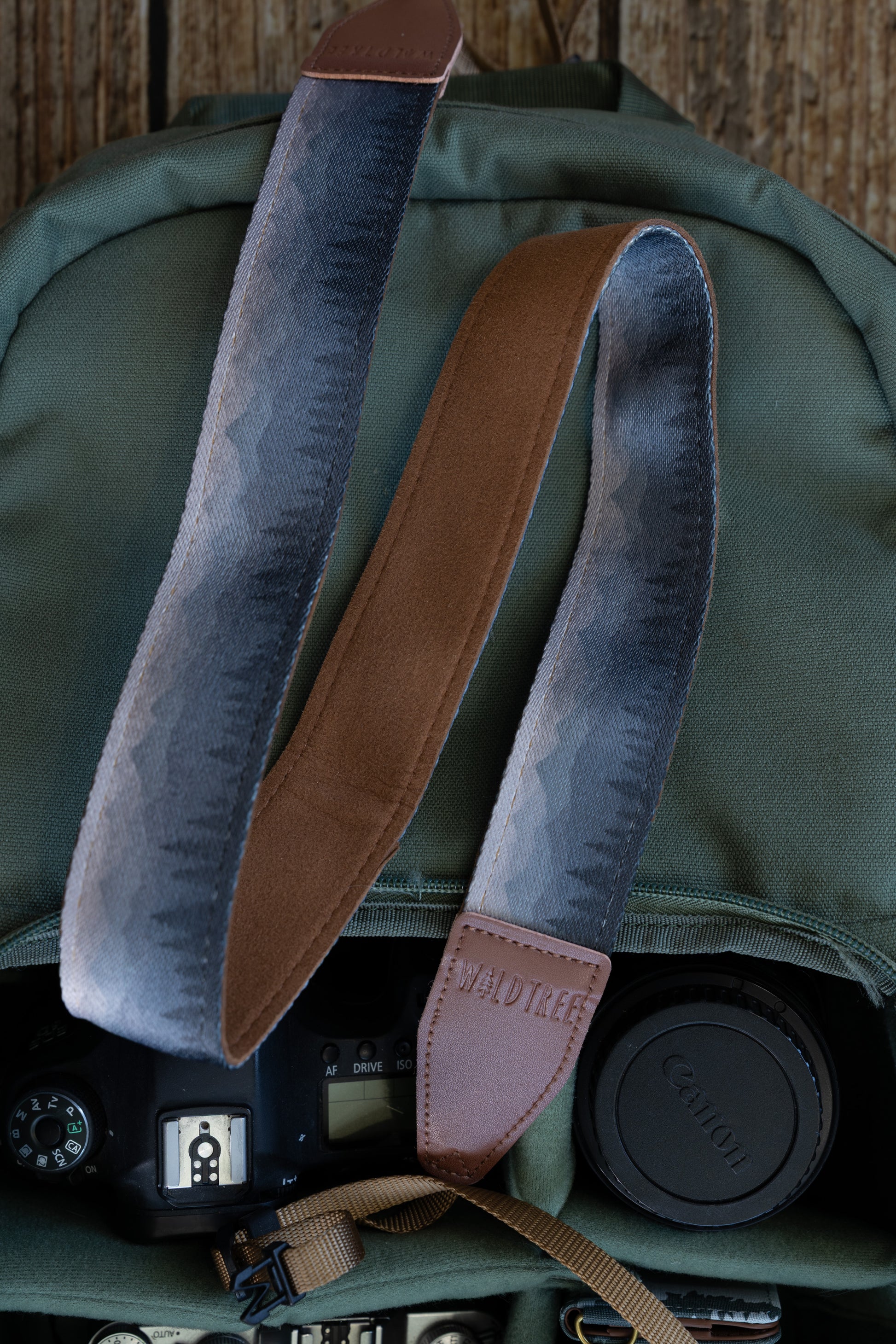 Silent Summit camera strap showcasing the dual-sided design with a blue-to-gray fading mountain silhouette print on the front and solid brown backing, resting inside an olive green backpack.