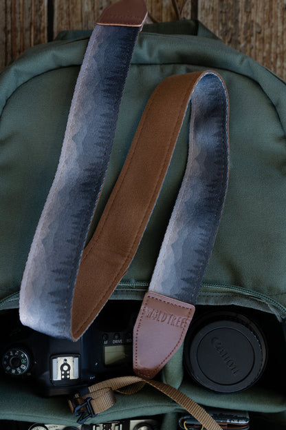 Silent Summit camera strap showcasing the dual-sided design with a blue-to-gray fading mountain silhouette print on the front and solid brown backing, resting inside an olive green backpack.