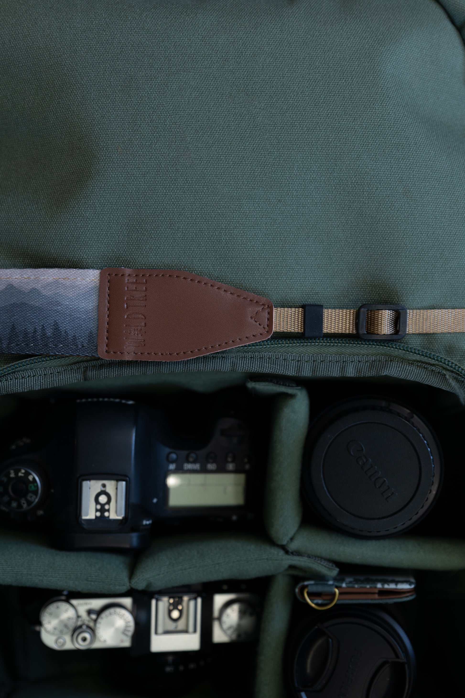 Silent Summit camera strap close-up of the spade-shaped brown leather connector piece with neat stitching and embossed Wildtree branding logo attached to tan nylon webbing.