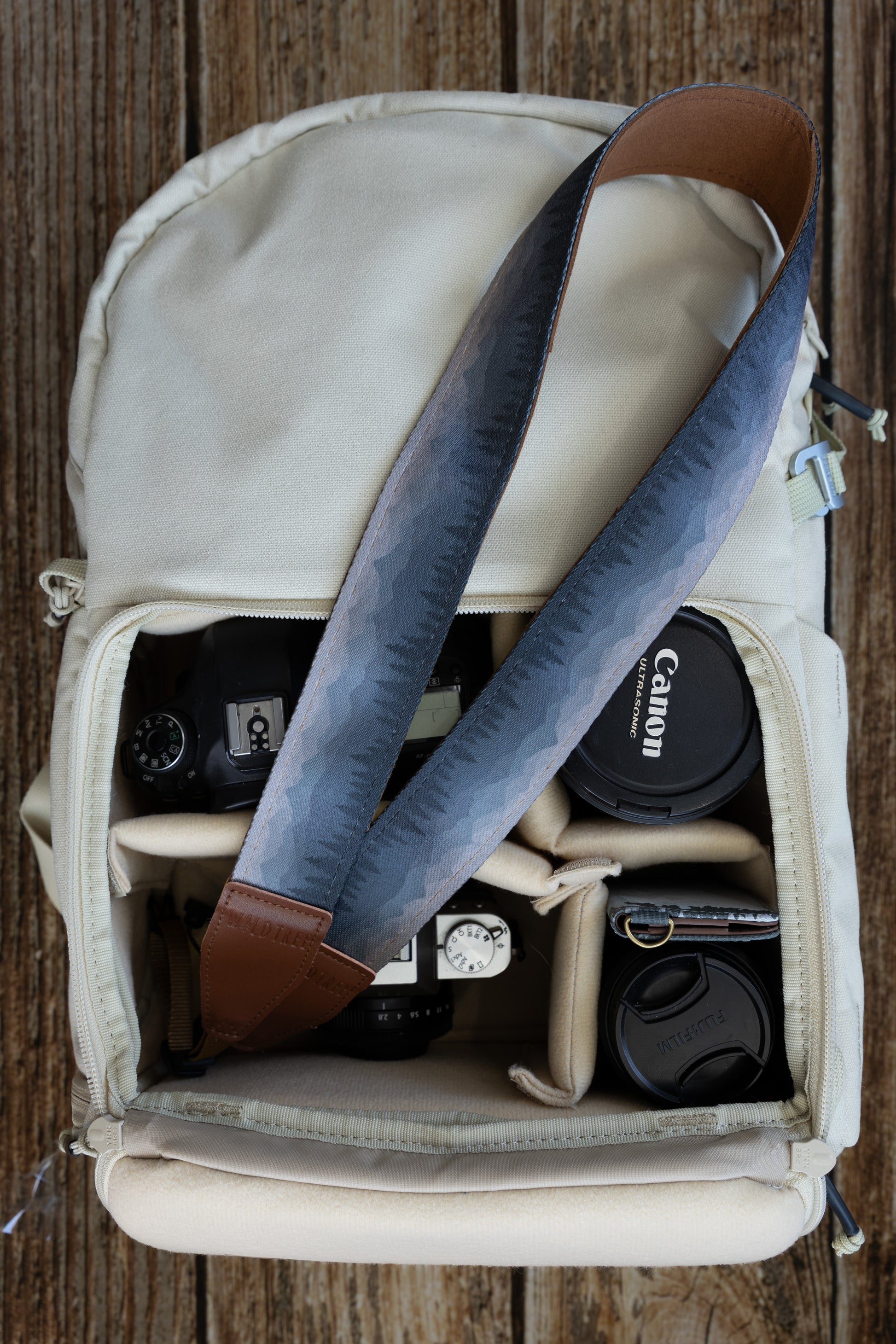 Silent Summit camera strap featuring a blue-to-gray gradient mountain range and pine forest pattern with a cognac brown underside, displayed over an open cream canvas camera backpack.