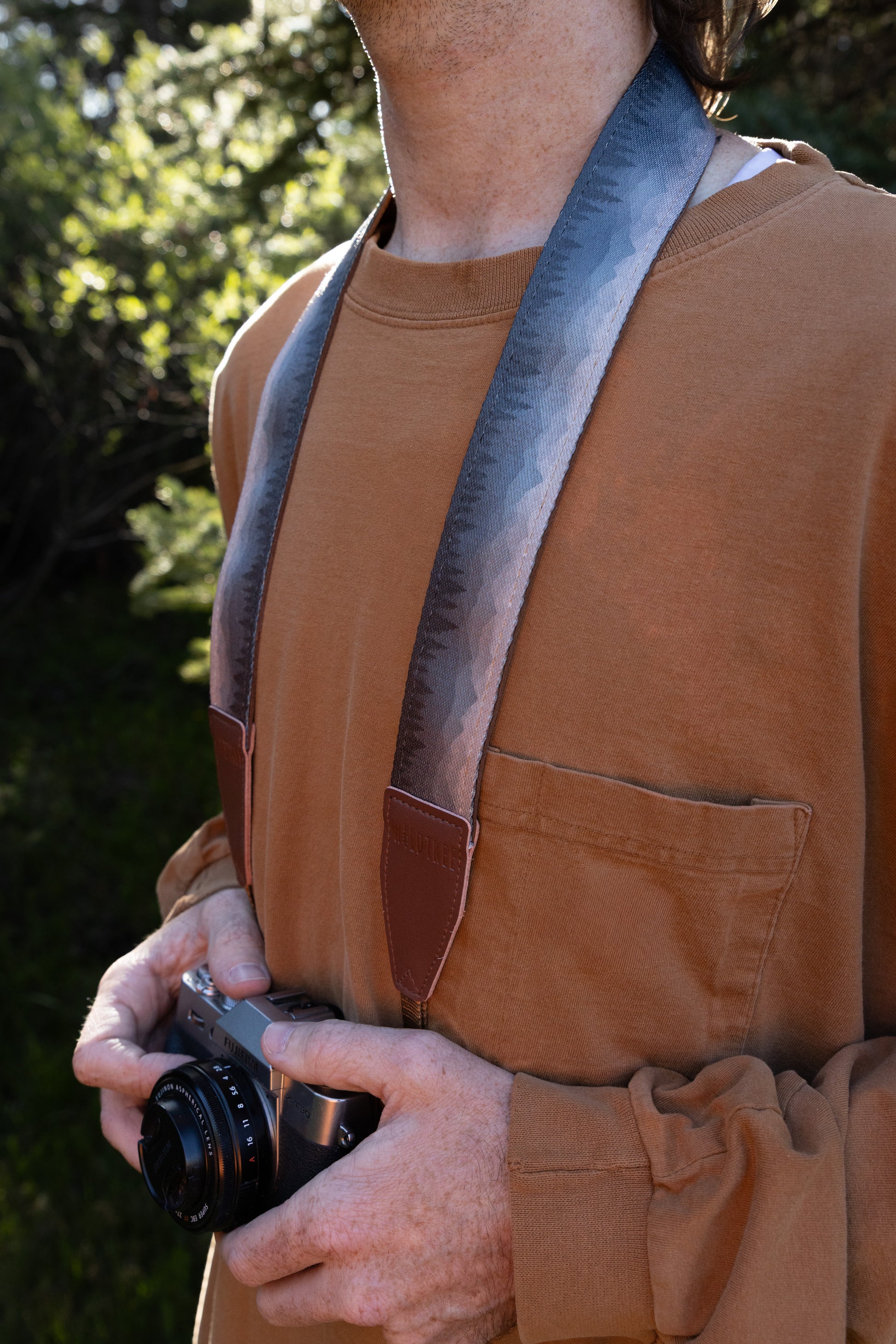 Silent Summit camera strap profile shot showing the subtle blue-to-gray wilderness landscape print draped over the shoulder of a photographer wearing an orange shirt.