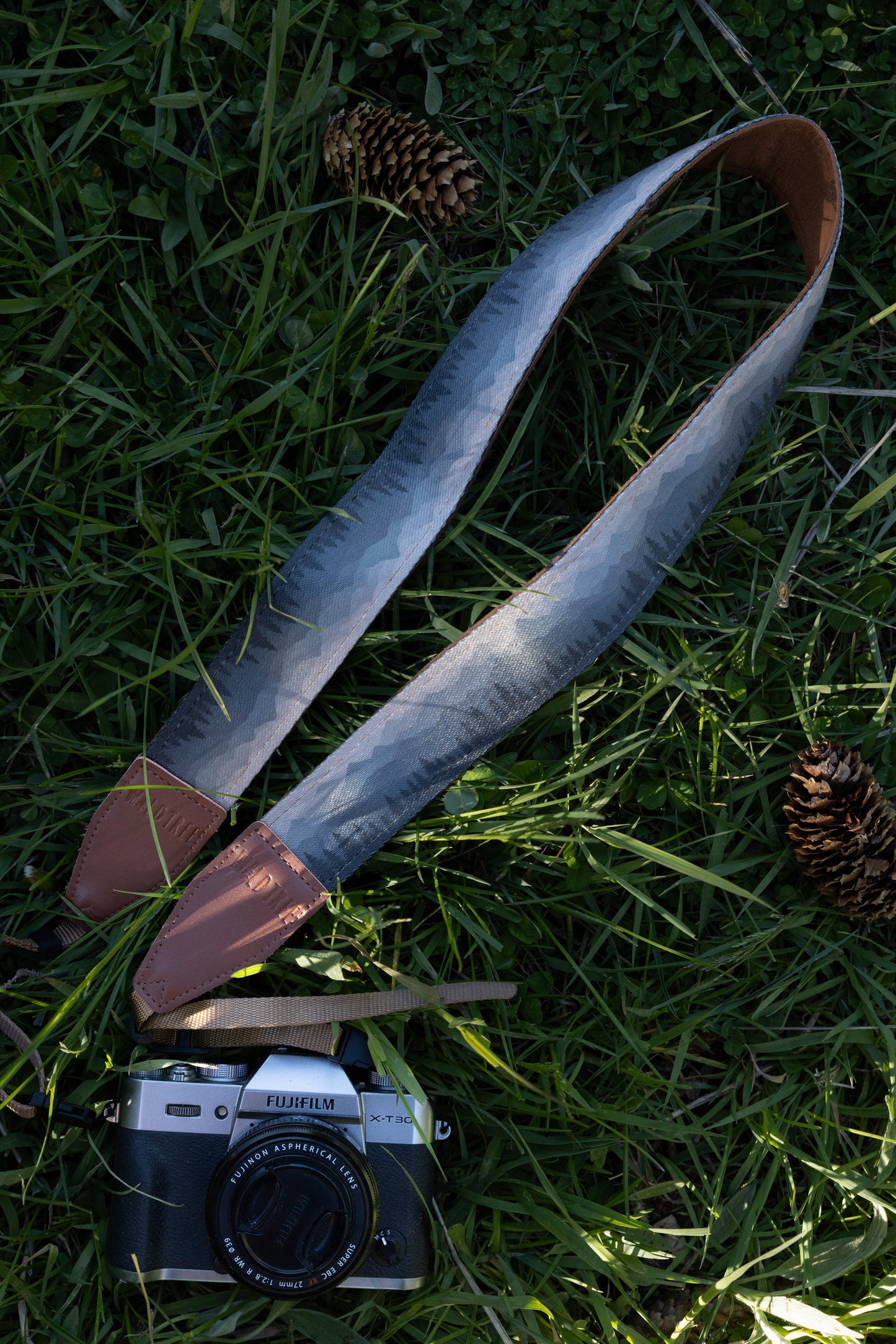 Silent Summit camera strap overhead full view displaying the repeating blue-to-gray gradient mountain range illustration, cognac brown accents, and adjustable tan camera loops on a grassy background.