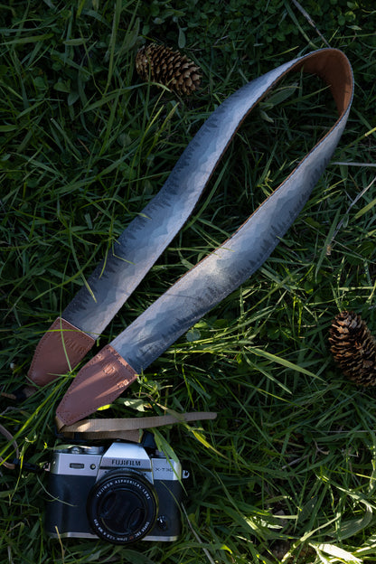 Silent Summit camera strap overhead full view displaying the repeating blue-to-gray gradient mountain range illustration, cognac brown accents, and adjustable tan camera loops on a grassy background.