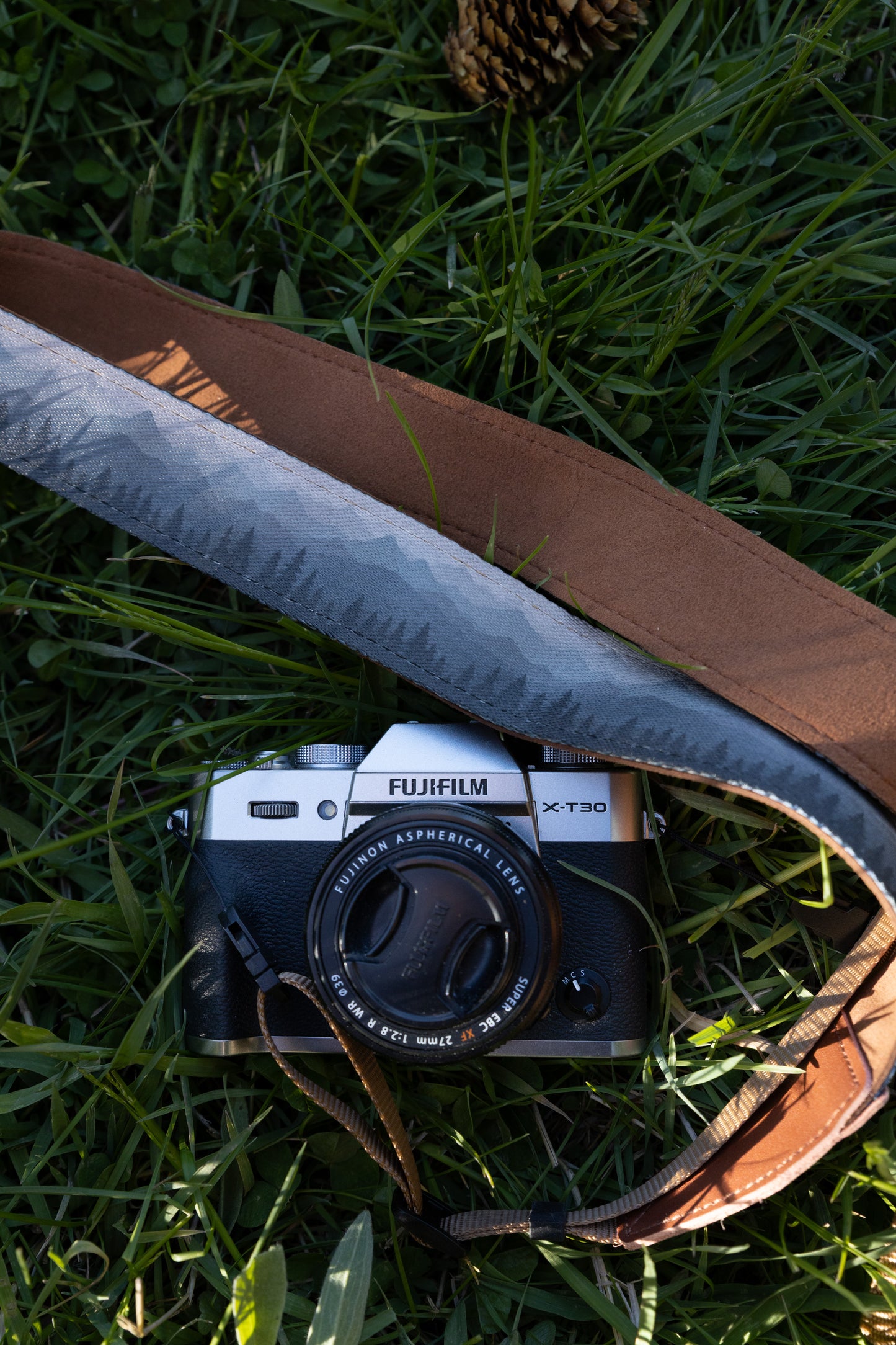 Silent Summit camera strap highlighting its distinct blue-to-gray ombre alpine landscape print and rich brown leather detailing, laying across a silver Fujifilm mirrorless camera in green grass.