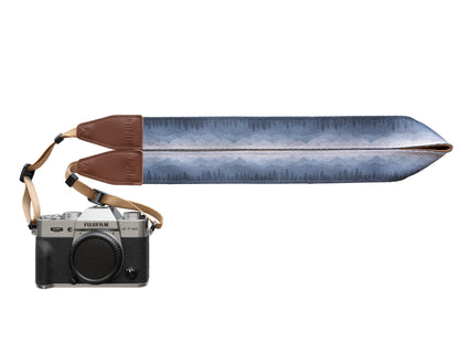 Silent Summit camera strap studio product design shot showing the folded fabric band with a blue-to-gray pine forest gradient illustration and stitched brown leather tips attached to a silver mirrorless camera.