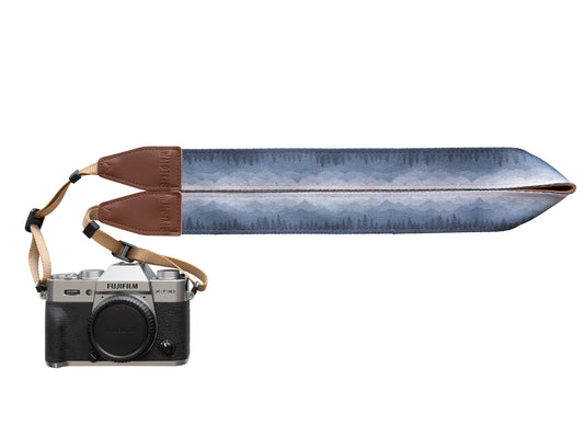 Silent Summit camera strap studio product design shot showing the folded fabric band with a blue-to-gray pine forest gradient illustration and stitched brown leather tips attached to a silver mirrorless camera.