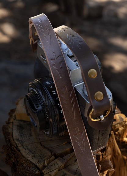 A vintage-style slim brown leather camera strap with hand-stamped evergreen tree patterns, attached to a retro-style silver mirrorless camera on a rustic wood stump.