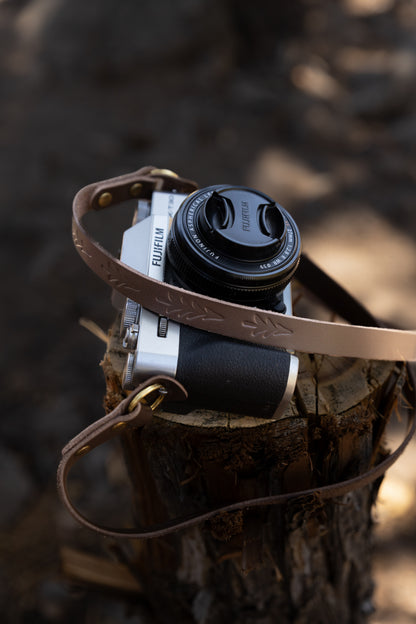 The Wildwood slim brown leather camera strap draped elegantly over a silver mirrorless camera resting on a weathered wood stump in soft outdoor light.