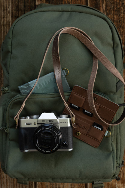 A vintage slim leather camera strap attached to a silver Fujifilm X-T30 camera, resting on a green outdoor backpack alongside a brown leather SD card wallet.