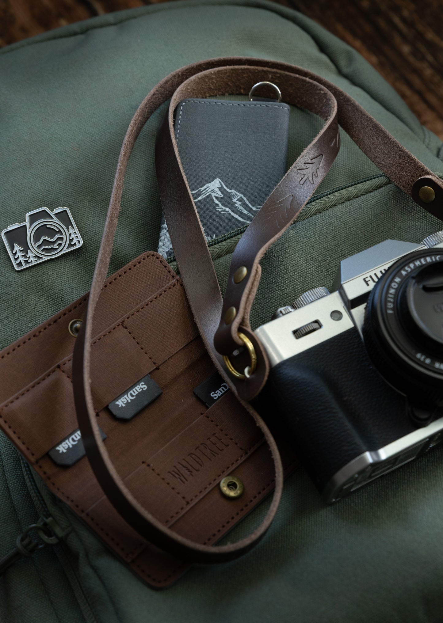 Overhead shot of the slim brown leather camera strap, a silver Fujifilm camera, and a brown leather SD card wallet on a green canvas backpack with an outdoor camera pin.