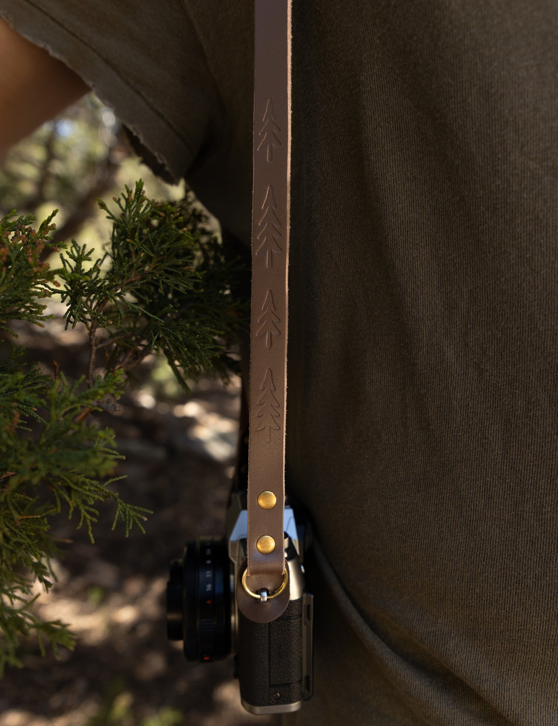 Detailed view of the vintage slim leather camera strap hanging vertically next to a green pine branch, showing the repeated hand-stamped tree pattern and bronze hardware.