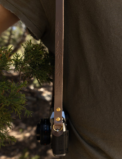 Detailed view of the vintage slim leather camera strap hanging vertically next to a green pine branch, showing the repeated hand-stamped tree pattern and bronze hardware.