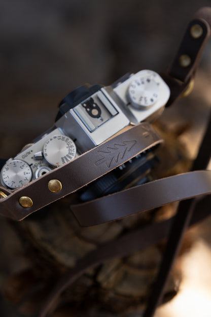 Close-up top view of the slim brown genuine leather strap resting on a silver camera body, highlighting the hand-stamped evergreen tree design and antique brass rivets.