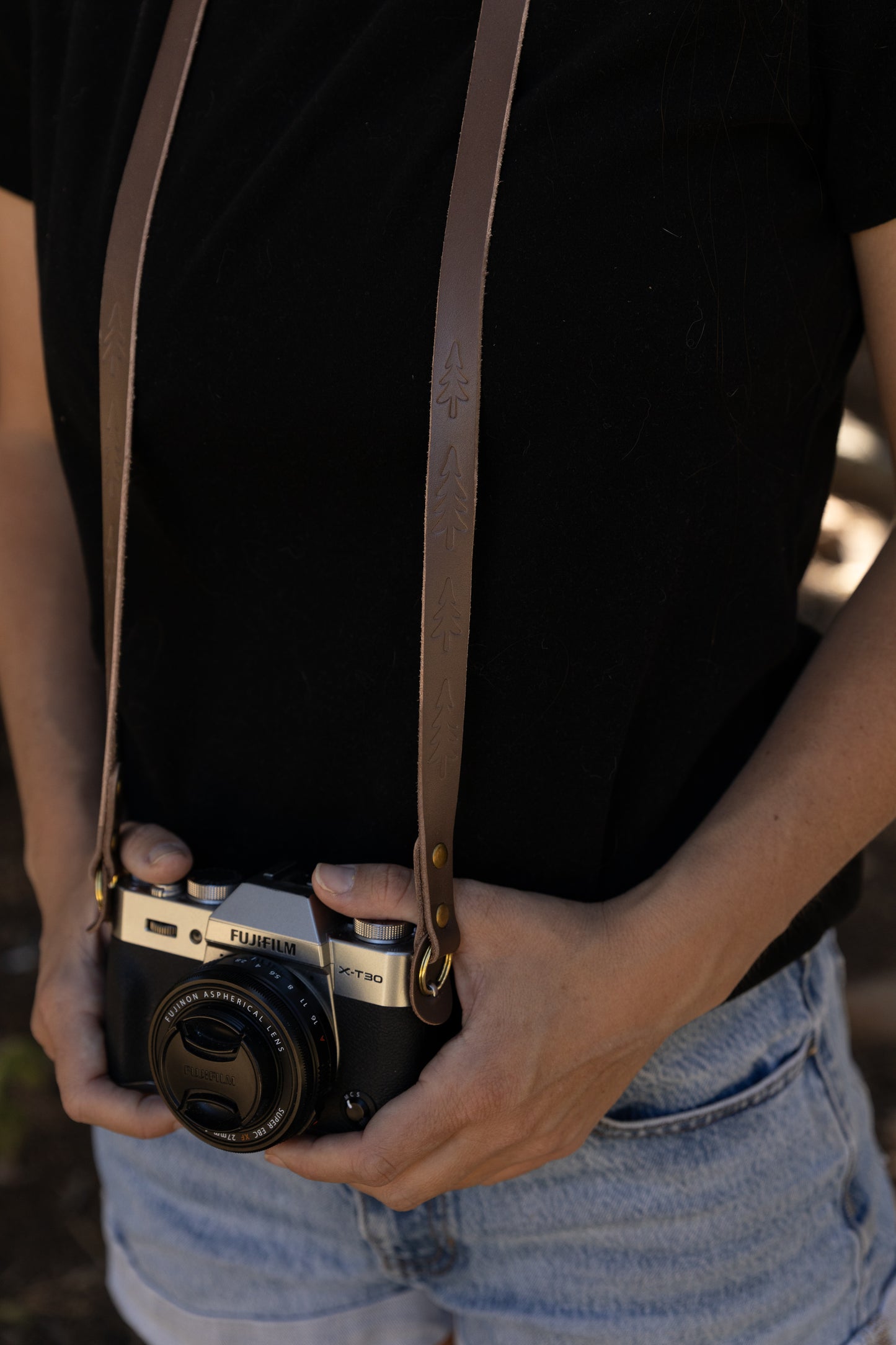 A photographer wearing the Wildwood vintage slim leather camera strap around their neck, holding a silver Fujifilm mirrorless camera during an outdoor shoot.