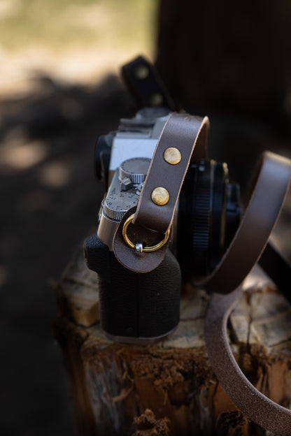 Detailed side view of the leather camera strap connection point, showcasing the premium antique bronze O-ring and the integrated leather scratch protector flap on a camera body.