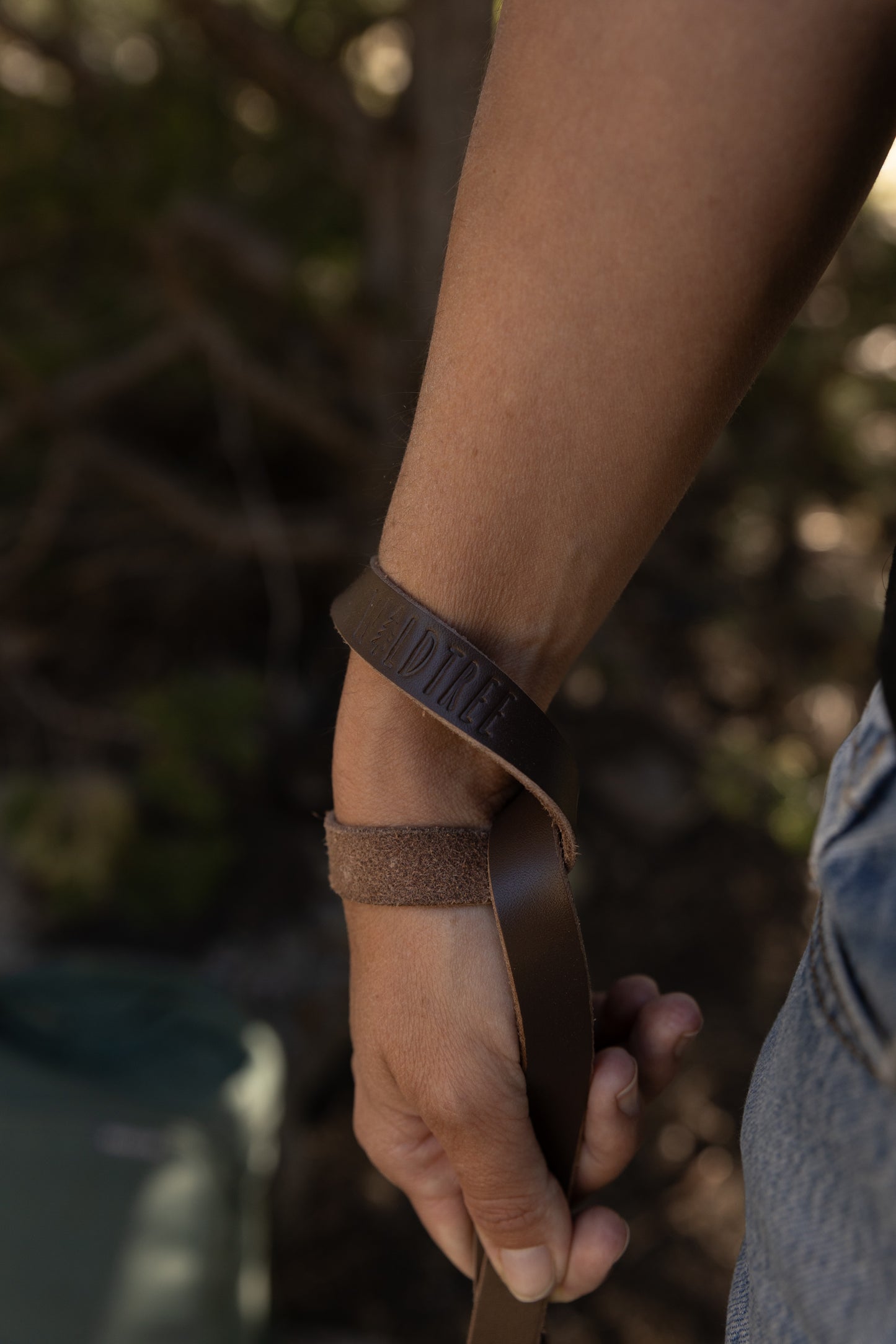 Close-up of the slim genuine leather camera strap wrapped around a person's wrist, highlighting the debossed Wildtree brand logo in the center of the leather.