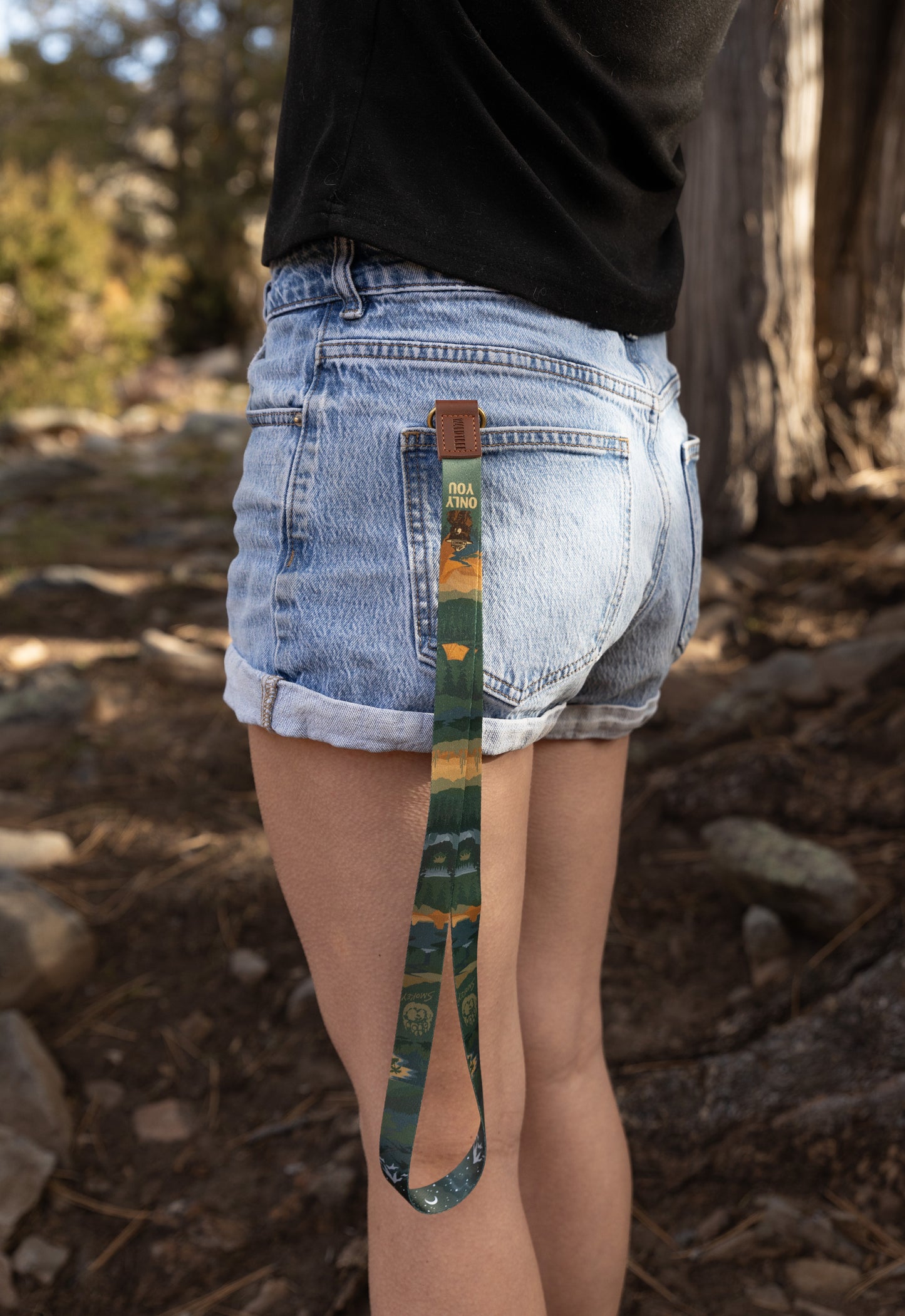 Back view of a person wearing denim shorts with the Smokey Bear neck lanyard hanging from a back pocket, showing the forest landscape design against an outdoor woodland background.