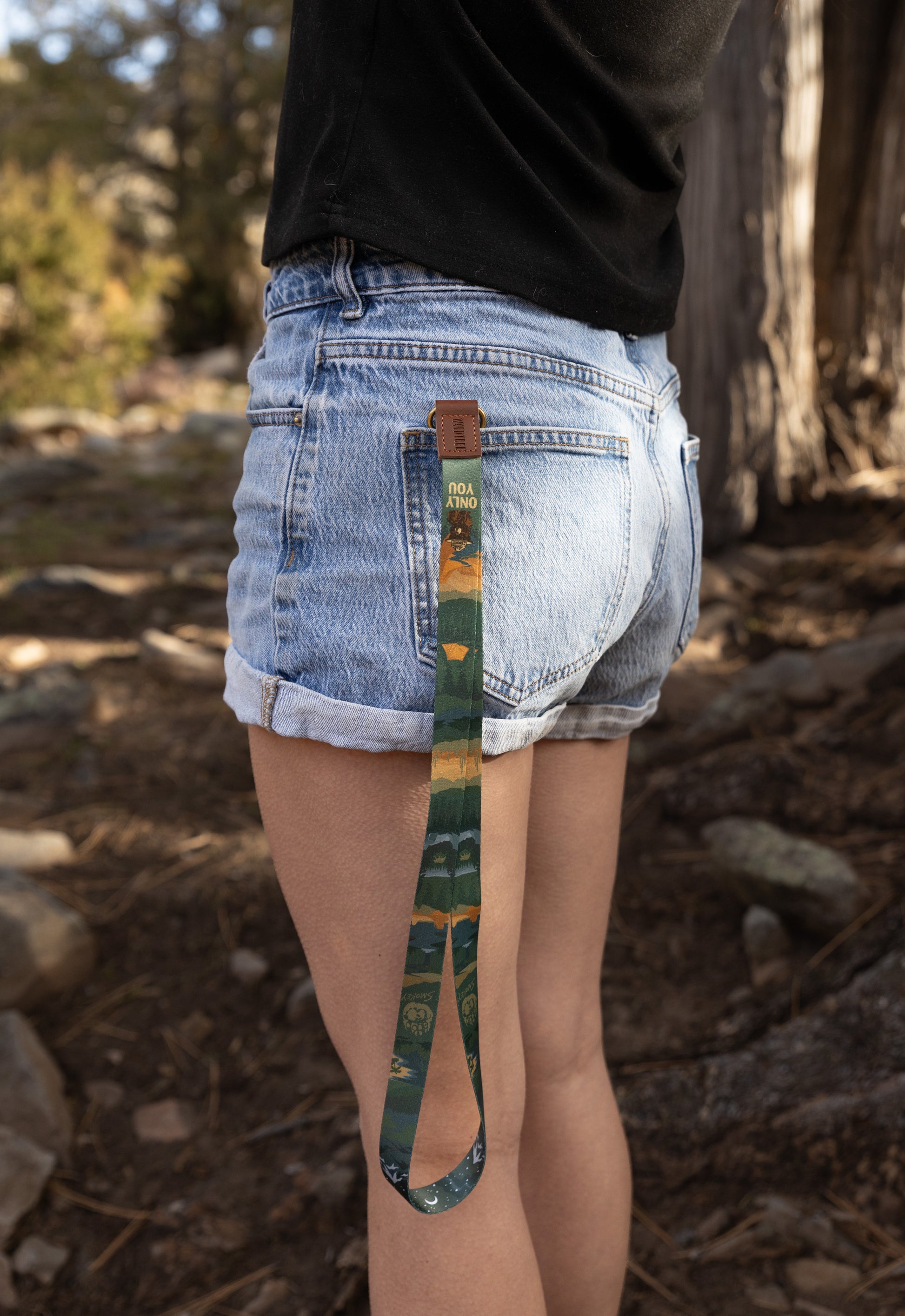 Back view of a person wearing denim shorts with the Smokey Bear neck lanyard hanging from a back pocket, showing the forest landscape design against an outdoor woodland background.