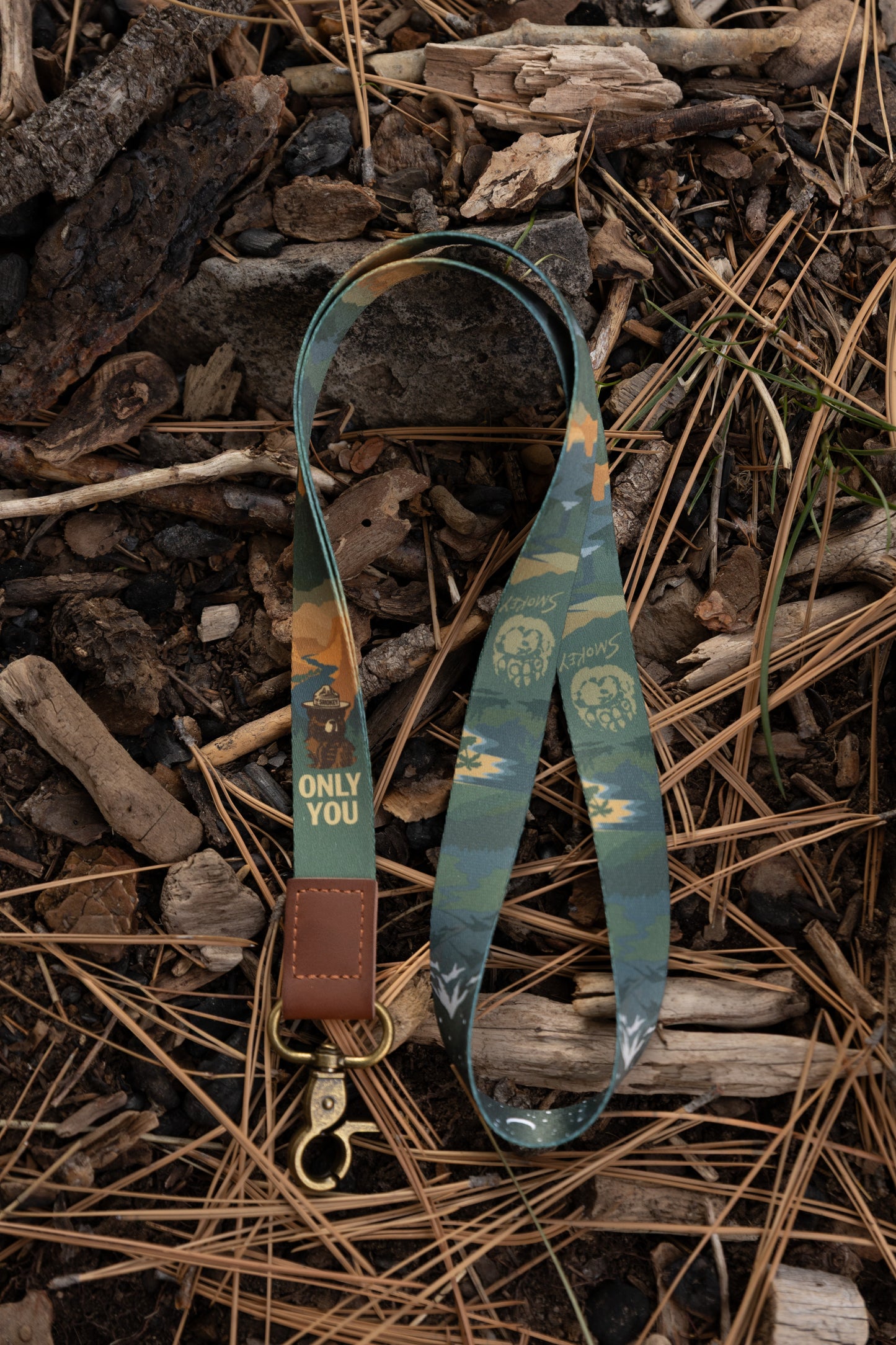 High-angle shot of the Smokey Bear lanyard lying on a forest floor covered in pine needles and twigs, highlighting the "Only You" text and the antique brass swivel lobster claw clasp.