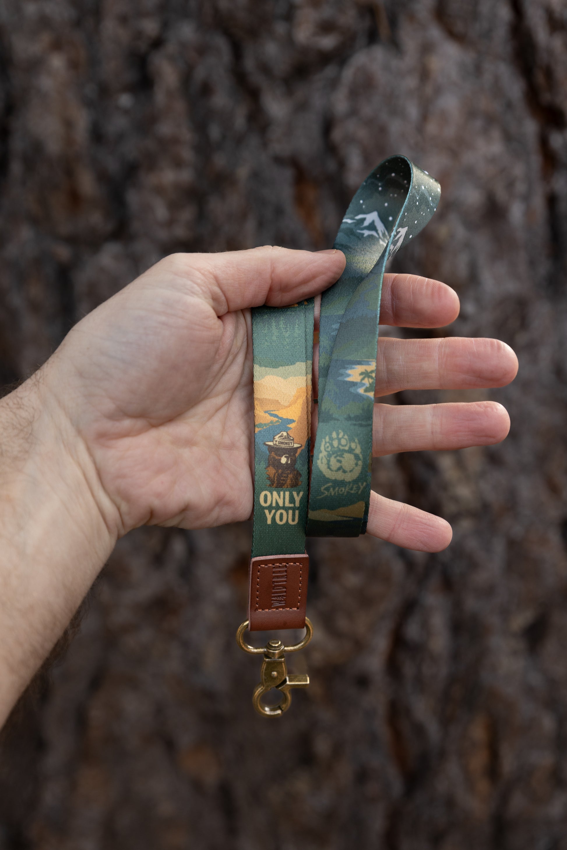 A person holding the Smokey Bear lanyard in their hand against a tree bark background, showing the full graphic of Smokey Bear and the night sky transition on the fabric.