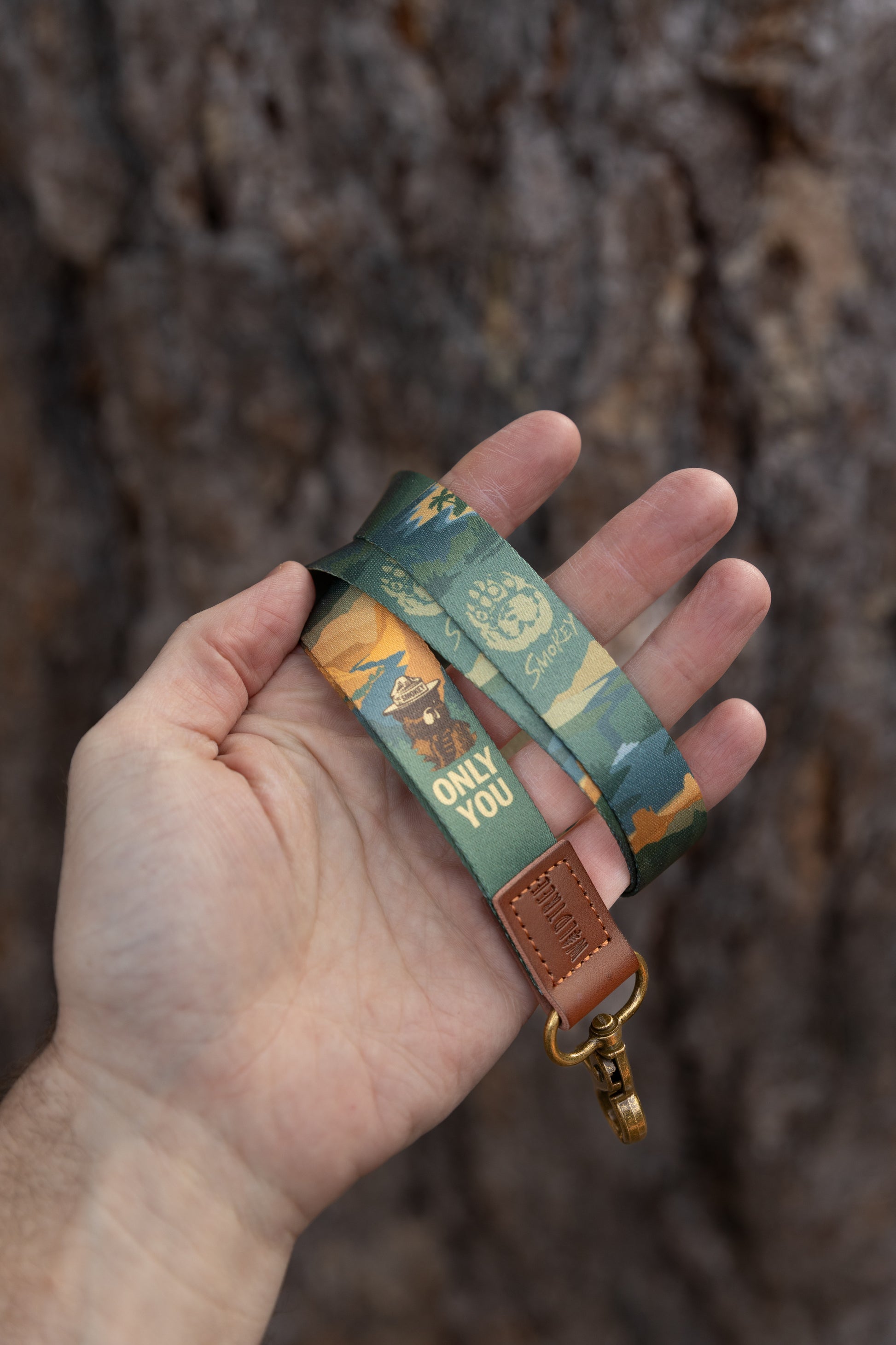 Close-up of the lanyard held in a hand, focusing on the debossed Wildtree logo on the brown leather attachment and the detailed mountain and paw print illustrations.