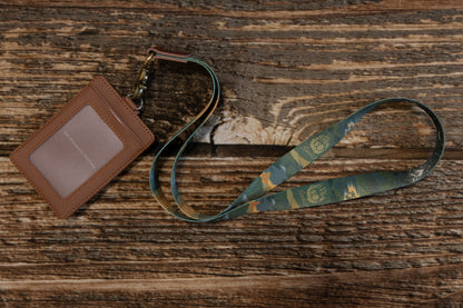 Wide shot of the lanyard and a brown ID card sleeve on a wooden tabletop, emphasizing the vintage aesthetic and the durable antique brass hardware.