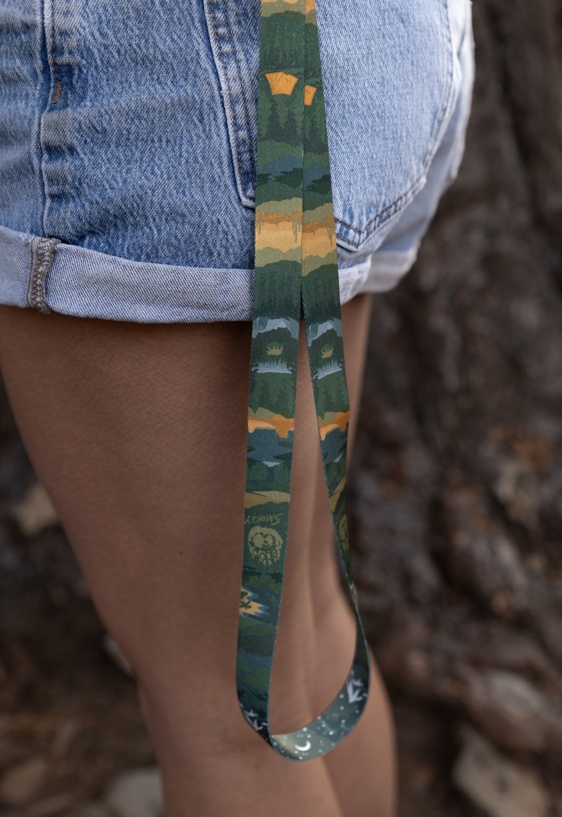 Extreme close-up of the lanyard hanging from a person’s pocket, showcasing the vibrant green and gold tones of the forest and mountain print.
