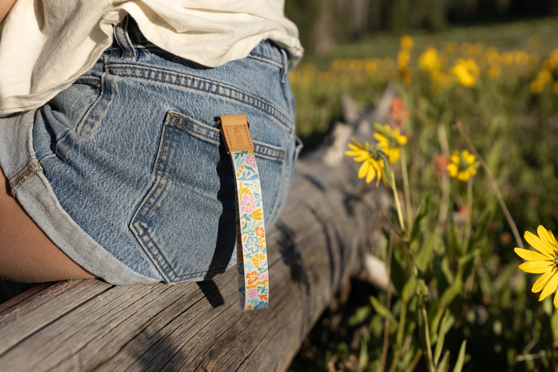 A person sitting outdoors on a log, wearing denim shorts. The colorful floral wristlet keychain hangs from a belt loop on the back pocket. Yellow wildflowers and green foliage are in the blurry foreground and background.