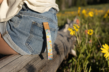 A person sitting outdoors on a log, wearing denim shorts. The colorful floral wristlet keychain hangs from a belt loop on the back pocket. Yellow wildflowers and green foliage are in the blurry foreground and background.