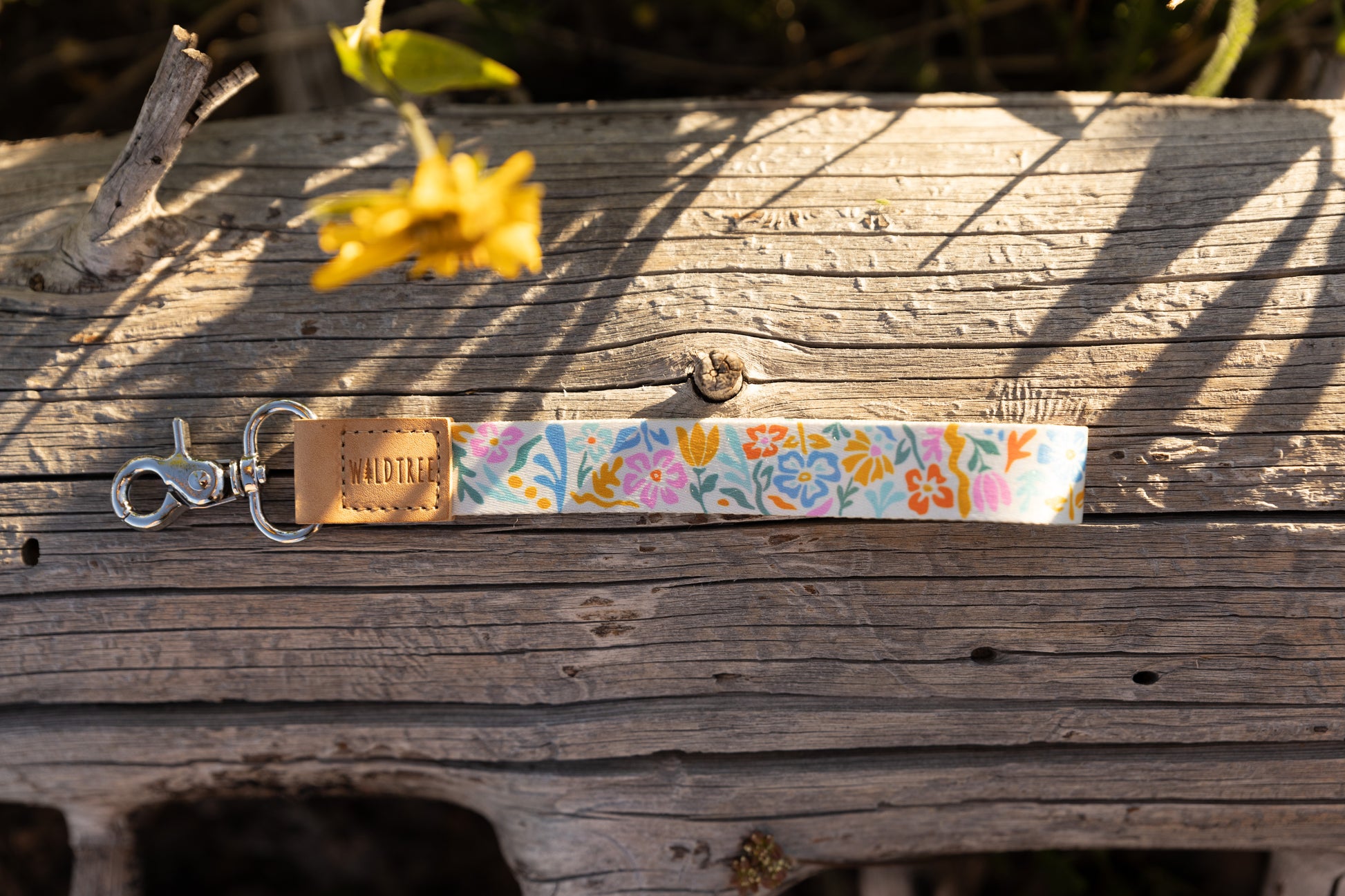A colorful wristlet keychain featuring a folk-art-style floral pattern in pink, blue, orange, and green on a white background. It is lying horizontally on a weathered gray log. The keychain has a light brown leather tag stamped with the "WILDTREE" logo and a silver lobster claw clasp. A blurred yellow flower is in the background.