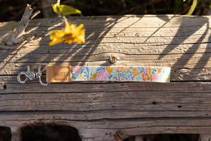 A colorful wristlet keychain featuring a folk-art-style floral pattern in pink, blue, orange, and green on a white background. It is lying horizontally on a weathered gray log. The keychain has a light brown leather tag stamped with the "WILDTREE" logo and a silver lobster claw clasp. A blurred yellow flower is in the background.