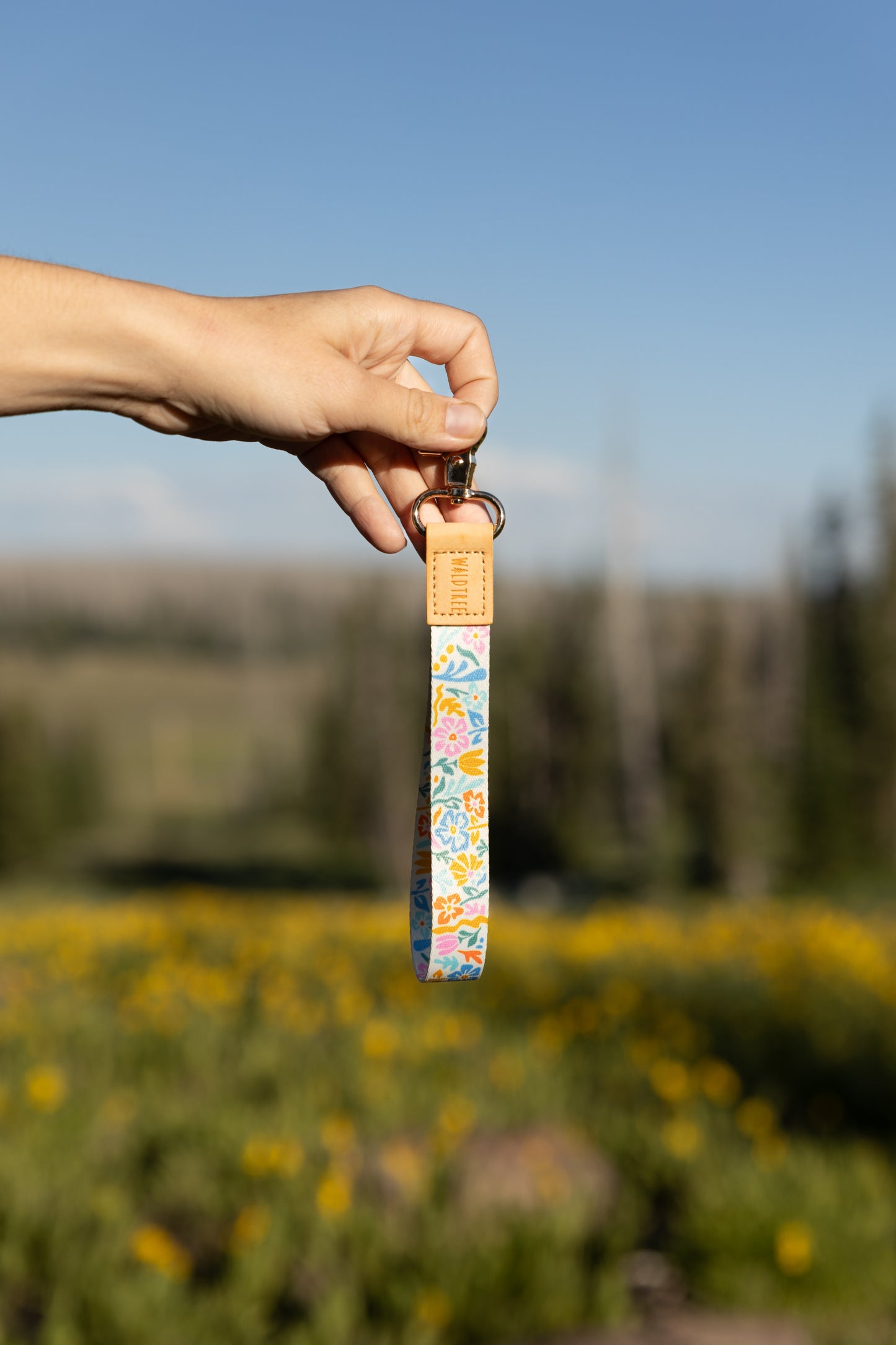 A person's hand holds the colorful floral wristlet keychain up against a clear blue sky and a blurred field of yellow wildflowers. The keychain has a light brown leather end and a silver clasp.
