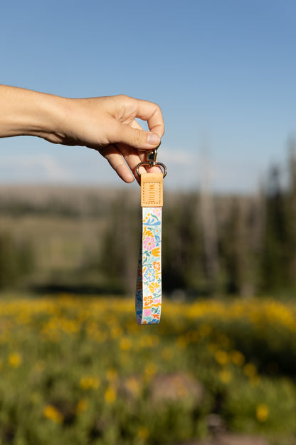 A person's hand holds the colorful floral wristlet keychain up against a clear blue sky and a blurred field of yellow wildflowers. The keychain has a light brown leather end and a silver clasp.