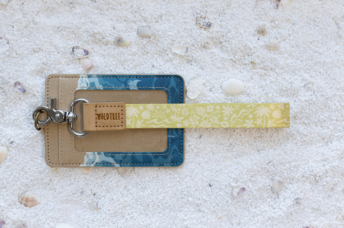 Sunny Day wristlet keychain clipped to the Ocean ID badge holder, both laid flat side by side, showing the badge holder's tan canvas top with pine tree emboss and teal shark-and-ray print body beside the cream-on-mustard beach and floral printed wristlet, on white sand with seashells.