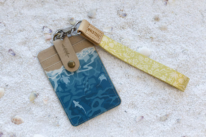 Close-up angled view of the Sunny Day wristlet keychain draped behind the Ocean ID badge holder, highlighting the tan leather Wildtree tabs, bronze snap closure, and teal shark-and-ray print body, on white sand with seashells.