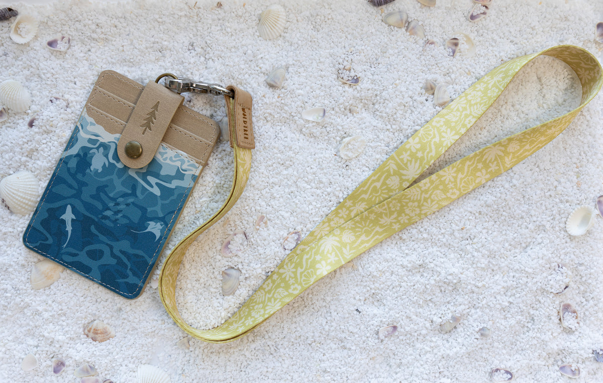 Sunny Day neck lanyard and Ocean ID badge holder arranged side by side, showing the cream-on-mustard beach and floral print alongside the badge holder's tan canvas and teal shark-and-ray print design, on white sand with seashells.