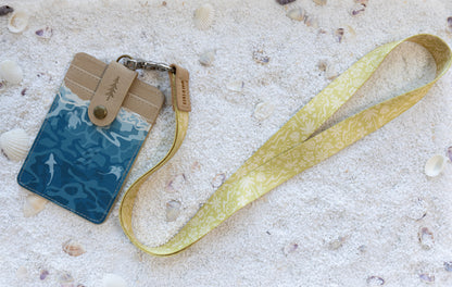 Sunny Day neck lanyard and Ocean ID badge holder arranged side by side, showing the cream-on-mustard beach and floral print alongside the badge holder's tan canvas and teal shark-and-ray print design, on white sand with seashells.