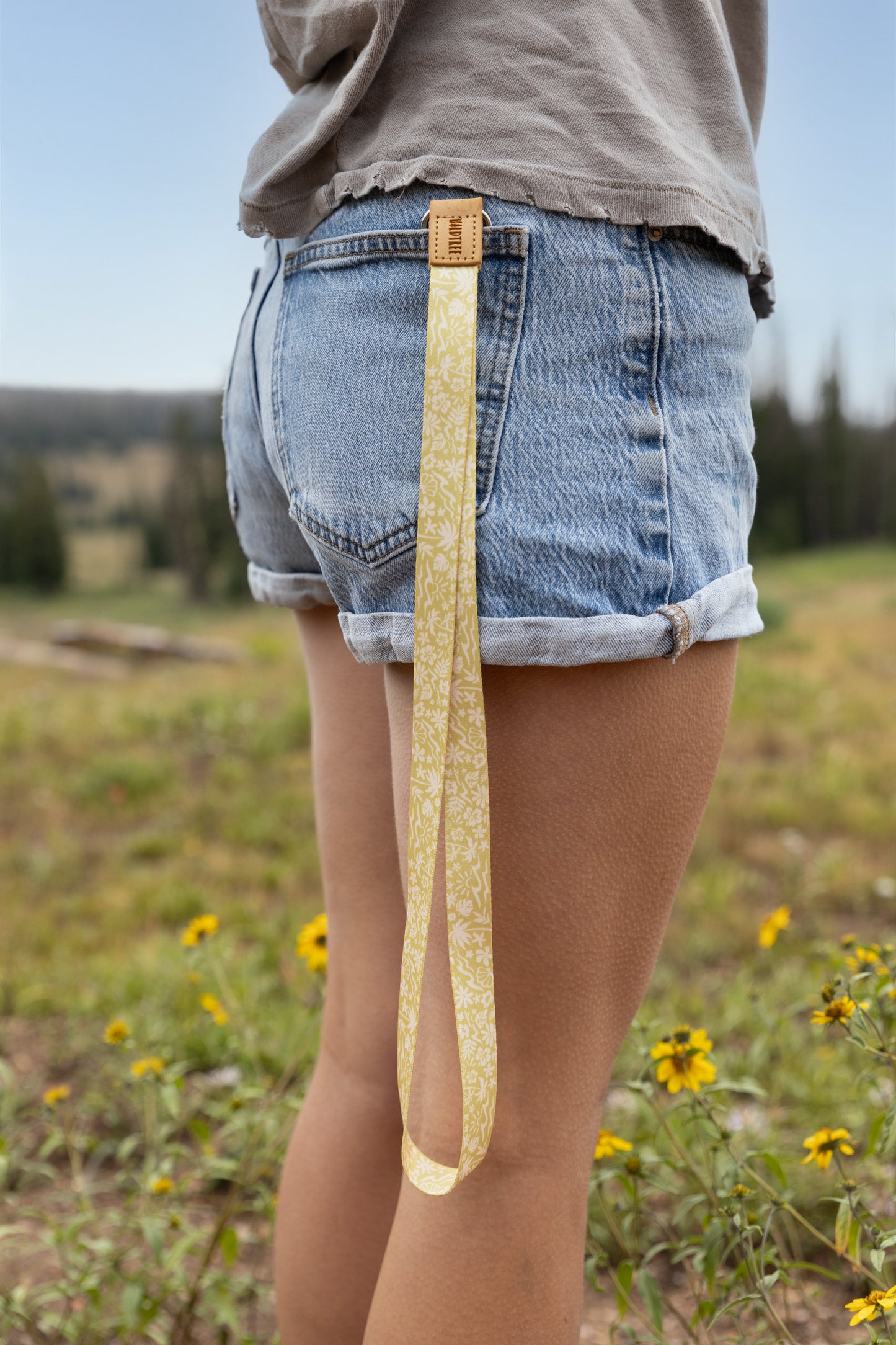 A person in cuffed denim shorts stands in a wildflower meadow under a blue sky with the Sunny Day lanyard clipped to their back belt loop, the yellow strap printed with waves, palm trees, suns, and seashells hanging down against their leg.