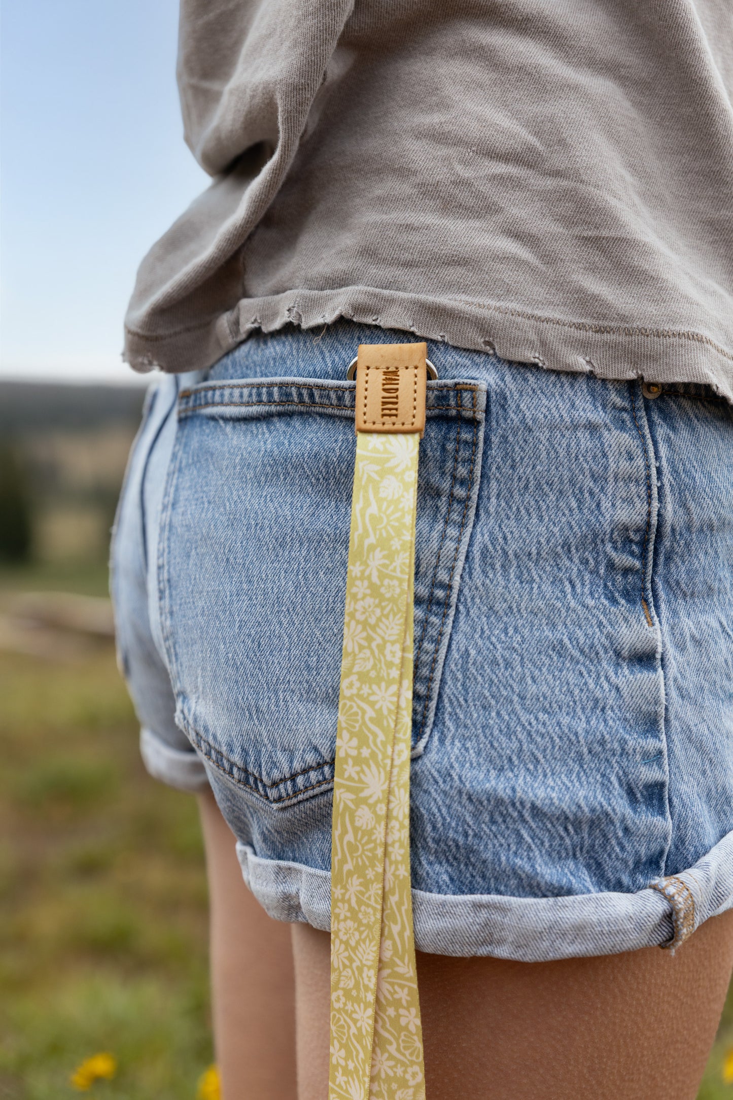 Close-up of the Sunny Day lanyard clipped to a denim shorts belt loop, showing the tan leather WILDTREE tab and the yellow strap printed with waves, palm trees, and seashells hanging against the wearer's leg in a grassy field.
