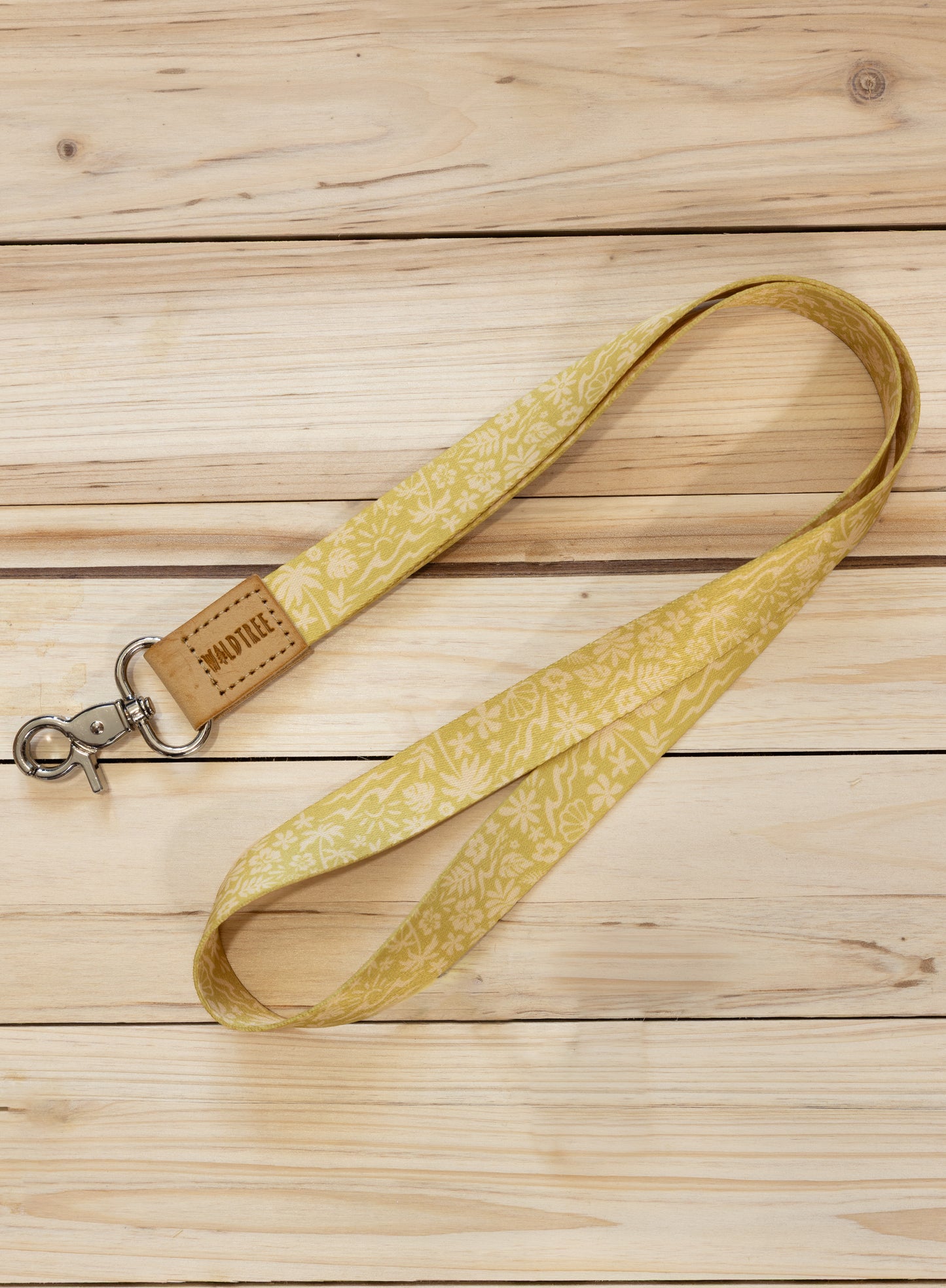 The Sunny Day neck lanyard laid diagonally across a light wood plank surface, showing the full yellow strap printed with a white beach-themed pattern of waves, palm trees, suns, flowers, and seashells, the tan leather WILDTREE tab, and the silver swivel clasp.