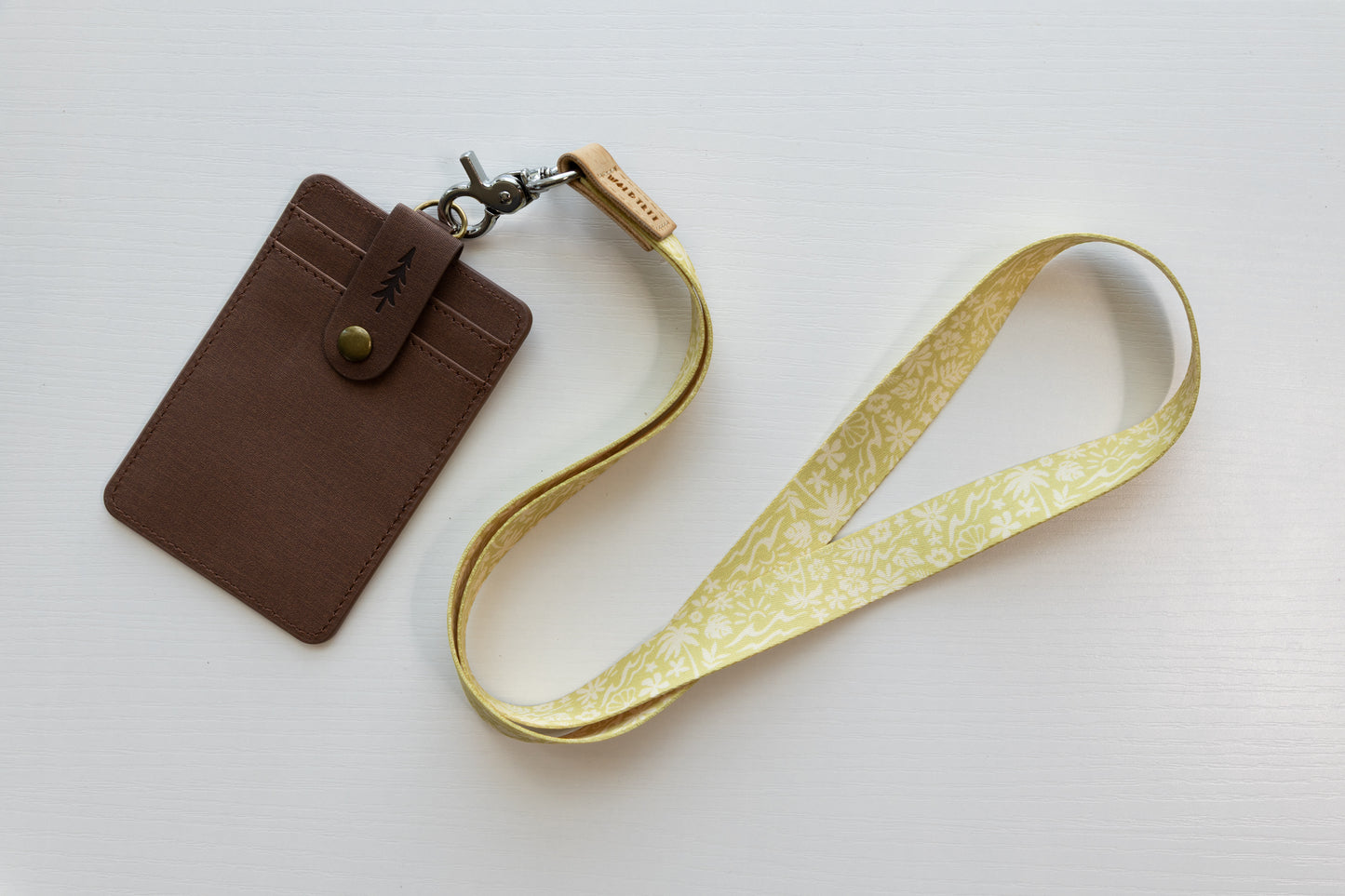 The Sunny Day neck lanyard on a white background paired with a brown vegan-leather card holder, connected by a tan leather WILDTREE tab and silver swivel clasp, the strap printed with a yellow and white beach pattern of waves, palm trees, suns, and seashells.
